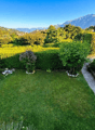 well-maintained garden with green lawn, hedges, and a gravel path, surrounded by trees and bushes, with a distant view of mountains