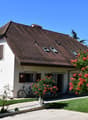 2 story house, white exterior, brown roof, multiple windows, bicycle parked in front, garden with red flowers