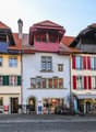 The image shows a row of traditional, colorful buildings with red tile roofs, wooden shutters, and decorative architectural elements. The buildings appear to be located in a town or city setting, with a cobblestone street in the foreground. The buildings