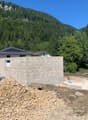 The image shows a house under construction in a mountainous, forested area. The foundation and walls of the house are visible, and there appears to be a garage or other structure next to the main building. The surrounding landscape is lush and green, with