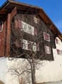 A building with a wooden wall, brick wall, and many windows. The house is surrounded by grass, and the snow is on the ground. It seems that the building is on the street. There is another building on the right, and it seems like there is a mountain in the