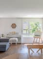 Spacious living room with large windows, grey sofa, wooden furniture, artwork, and potted plants. The room has a cozy and modern feel with a balcony or terrace visible through the windows.