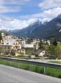 Mountain town with houses, mountains, road, cars, crane, trees, grass, sky