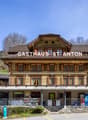 A traditional Swiss-style wooden building with a large balcony and terrace, housing a Gasthaus (inn or guesthouse) called 'Gasthaus St. Anton'. The building has a prominent sign displaying the name and is surrounded by trees and a grassy area.