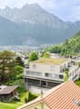 The image shows a picturesque mountain landscape with a small town nestled in the valley below. The town is surrounded by lush green forests and rolling hills, with snow-capped peaks in the distance. The buildings in the town appear to be a mix of traditi