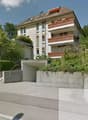 Multistory building, white walls, brown roof, wooden balconies, garage, concrete stairs, trees and shrubs