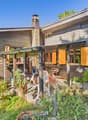 wood house, orange shutters, garden with plants, brick pathway, chimney, porch with table and chairs, grill, trees in the background