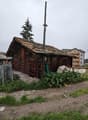 A wooden house with a rusty roof and a chimney, surrounded by plants and grass, with a large pole on its roof.