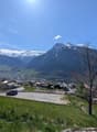 The image shows a scenic mountain landscape with snow-capped peaks, a small village nestled in the valley below, and a clear blue sky with a few clouds. There are solar panels visible on some of the buildings, indicating the use of renewable energy. The f