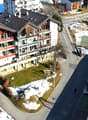 Multi-story apartment building with red and white exterior, balconies, surrounded by snow-covered landscape, with a road and ski lift visible in the background.