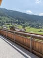 Wooden balcony with a view of a mountainous, forested landscape. The balcony has a table and chairs, and overlooks a village nestled in the valley below.
