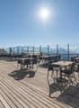 Outdoor dining area with wooden deck, tables and chairs, surrounded by glass railings offering a panoramic view of snow-capped mountains under a bright, sunny sky
