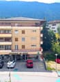 A multi-story residential building with a pharmacy on the ground floor, surrounded by trees and mountains in the background. The building has balconies and there are several cars parked in the lot in front of the building.