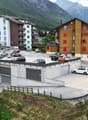 The image shows a residential area in a mountainous region. There are several multi-story apartment buildings and houses, some with colorful facades. There is a parking lot in the foreground with several cars parked. The surrounding landscape includes for