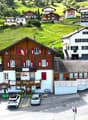 Mountain village complex with multiple buildings, wooden facades, and red shutters. One building has a restaurant sign. The complex is surrounded by a green hillside. Cars parked in front of the buildings.
