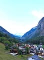 cluster of houses, green trees, rocky mountains, church, grassy fields
