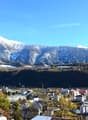 View of mountains with snow in background, surrounded by a town with various buildings, trees and greenery