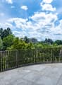 The image shows an outdoor terrace or balcony with a metal railing overlooking a scenic landscape. The terrace has a stone or concrete floor, and the view beyond the railing includes lush green trees, a building in the distance, and a partly cloudy blue s