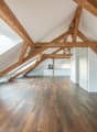 Spacious attic room with exposed wooden beams, hardwood flooring, and a skylight providing natural light. The room appears to be an open-concept living space with a kitchen visible in the background.