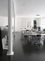 Large open office space with multiple desks, computers, and office equipment. The room has high ceilings, white walls, and dark hardwood floors. There is a dishwasher visible in the corner of the room.