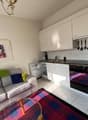 The image shows a studio apartment with an open-plan layout. The kitchen area features white cabinets, a built-in oven, and a tiled backsplash. The living area has a gray sofa with colorful throw pillows, a coffee table, and a framed artwork on the wall.