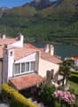 Large house with red roof, white walls, multiple windows, surrounded by greenery, mountains in the background, lake nearby