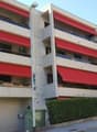 Multi-story apartment building with red and white exterior, balconies on each floor, and a parking area in front of the building.