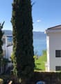 Two white buildings, one with a balcony, surrounded by greenery, with a lake and mountains in the background.