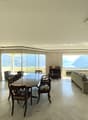 Spacious room with a dining table, chairs, a cabinet, a couch, an ottoman, a fireplace, and bookshelves. Large windows offer a scenic view.
