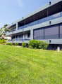 A modern gray building with large windows and a glass balcony. It has a spacious lawn with bushes and trees in front.