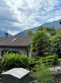 stone house with brown roof, stone walled exterior, solar panel on roof, surrounded by greenery, mountains in the background