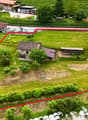 Empty plot of land with house in the middle, red line indicating boundary, surrounding are houses, cars parked, street light, trees, vineyard