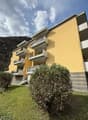 Yellow building with balconies on each floor, stairs leading up to building, small plants and bushes in front