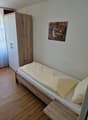 The image shows a bedroom with a wooden wardrobe, a bed with white bedding, and a framed artwork on the wall. The room has a hardwood floor and appears to be a simple, functional living space.
