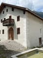 Large white building with brown roof, 2 floors, many windows, wooden door, wooden balcony, gravel path, fountain
