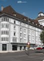 Multi-story building, white exterior, brown roof, many windows, shops on the ground floor, parking lot in front