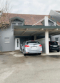 grey residential building with tiled roof, covered parking lot, two cars parked, two trash bins, tree, small stone wall