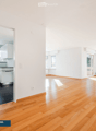 Open-plan space with wooden flooring, white walls, and large windows. The area includes a kitchen section with cabinets and a sink, and a living area with ample natural light.