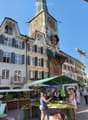 Town square with buildings, large clock tower, green market umbrellas, plants for sale