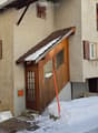 Two story house with wooden front door, brick wall, wooden window shutters, snow covered roof and yard