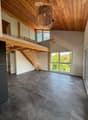 Open-plan living space with exposed wooden beams, grey tiled floor, large windows, and a wooden staircase leading to an upper level.