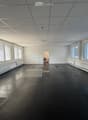 Empty room with white walls, black floor, large windows with blinds, and a doorway leading to another room.