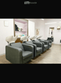 A salon with a row of grey chairs, a wooden floor, a white wall, a shelf with products, and a mirror.
