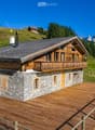 A two-story wooden chalet with stone base, large balcony, and wooden deck, surrounded by green hills and mountains.