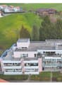 Aerial view of a modern apartment building with multiple balconies, surrounded by a green field and trees, with a clear view of the surrounding landscape.