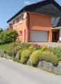 Two-story house with orange exterior, white garage door, satellite dish, and well-maintained garden with colorful flowers and shrubs.