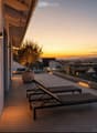 Modern rooftop terrace with sun loungers, a small table, and a potted tree, overlooking a cityscape at sunset.