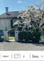 A two-story house with a tiled roof, white exterior walls, and black shutters. The house is surrounded by a garden with a variety of plants and trees, including a cherry blossom tree in full bloom. The driveway is paved with gravel, and there is a black g