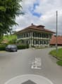 A street view of a house with a car parked in front. The house has a brown roof and white walls with green shutters. The house is surrounded by green grass and bushes. There are other houses in the distance, and the sky is cloudy.