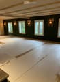 Empty room with dark green walls, wooden floor, and several windows. Exposed wooden beams on the ceiling and wall-mounted lamps are visible.